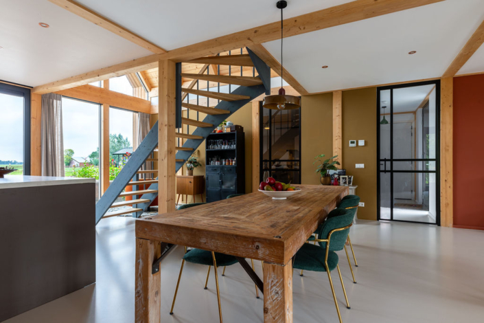 Moderne eetkamer met grote houten tafel en zes groene stoelen, wenteltrap naar boven. Grote ramen en glazen deur laten natuurlijk licht binnenstromen. Kom met fruit op tafel nodigt uit. - Finnhouse
