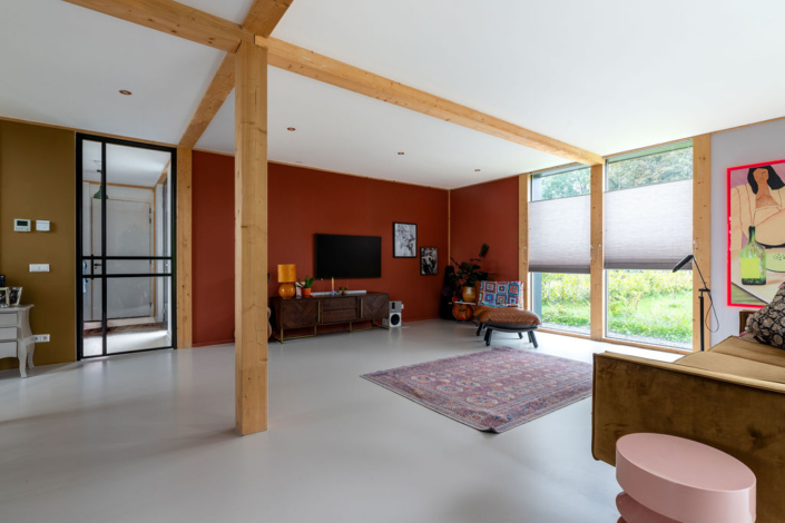 Gezellige woonkamer van Finnhouse met houten balken, roestkleurige muren en grote ramen. Comfortabele bank, tv, fauteuil met gitaar, kleurrijke tapijt en schattig aangeklede decoratie. Uitzicht op prachtige tuin. - Finnhouse