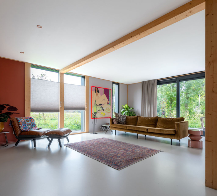Moderne woonkamer van Finnhouse met bruine fluwelen bank, houten balken en grote ramen. Twee leren stoelen, een kleurrijke tapijt en kunst zorgen voor een uitnodigende sfeer. Minimalistisch en ruimtelijk ontwerp. - Finnhouse