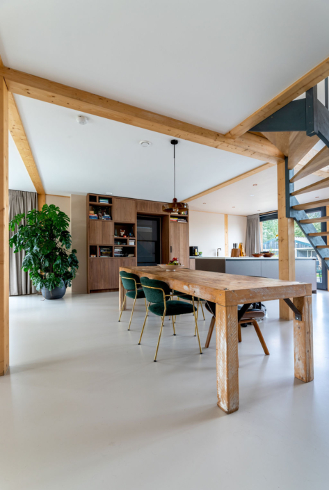 Gezellige moderne eetkamer geschikt voor een gezin, grote houten tafel met groene stoelen omringd door plant en open schappen met decor. Rechts trap, grote ramen zorgen voor veel natuurlijk licht. - Finnhouse