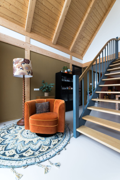 Gezellige woonkamer ideaal voor een gezin, met een oranje fauteuil op een patroonkleed, houten trap met zwart hekwerk en donker kastje met plant onder houten gewelfd plafond. - Finnhouse