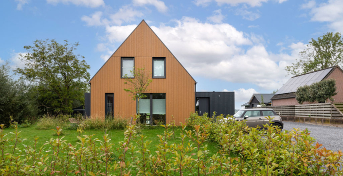 Modern driehoekig huis met houten gevel, grote ramen en puntdak, achtertuin door groen landschap. Kleine auto op grindoprit rechts. Perfect voor een gezin; ander huis met zonnepanelen op achtergrond. - Finnhouse