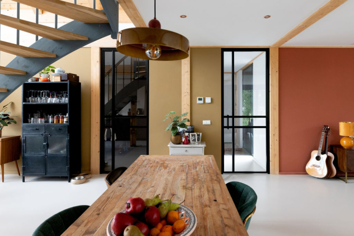 Gezellige eethoek met houten tafel, groene stoelen en fruitschaal als middelpunt. Gitaar tegen de muur, glazen deuren, metalen kast met glaswerk en trap met houten details in moderne gezinswoning. - Finnhouse