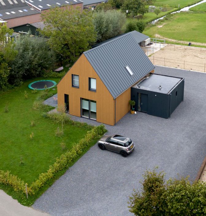 Luchtfoto van een moderne houten woning met donkere dakbedekking, grote ramen en aangebouwde garage. Gelegen op een groen grasveld met trampoline, ideaal voor gezinnen, achtertuin door velden en bomen. - Finnhouse
