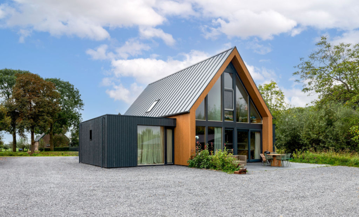 Modern A-frame huis met grote ramen en metalen dak, omgeven door bomen. Donkere houten gevelbekleding en een knusse patio met stoelen en tafel, ideale gezinsuitjes. Gelegen aan een grindpad. - Finnhouse