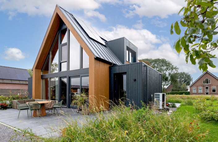 Moderne houten Finnhouse met zwarte panelen, grote ramen en zonnepanelen op het dak. Voorgrond toont patio met tuinmeubilair, binnenplaats groen onder een deels bewolkte hemel. Perfect voor een gezin. - Finnhouse