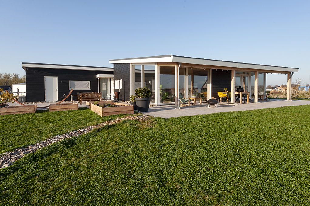 Moderne ecologische Finnhouse woning met zwarte gevel, grote ramen en ruime veranda. Voortuin heeft groen gras, stenen pad, bloembakken en zitgedeelte. Heldere blauwe lucht horizontale duurzame bouwstijl. - Finnhouse