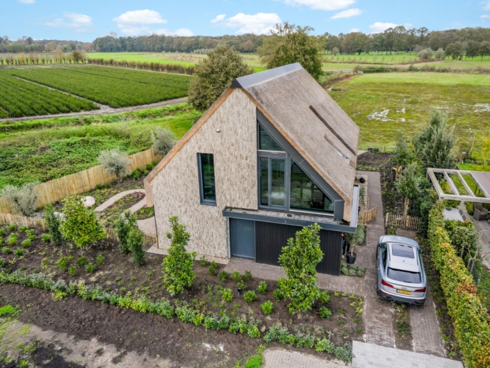 Luchtfoto van een modern huis met driehoekig, rieten dak, ideaal voor een gezin. Omringd door aangrenzende tuin en velden; auto geparkeerd op rit. Bomen en groen reiken ver onder deels bewolkte hemel. - Finnhouse