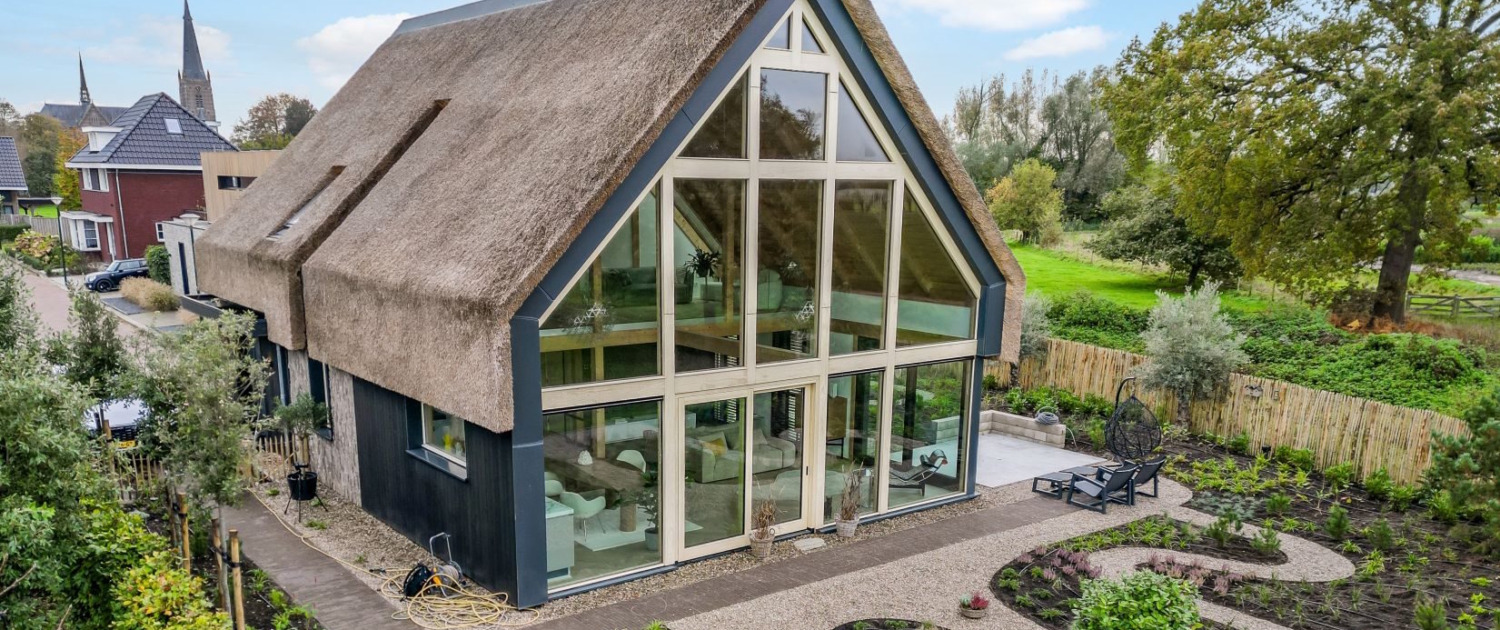 Moderne Finnhouse met rieten dak, grote glazen ramen, omgeven door aangelegde tuinen en paden. Op de achtergrond verrijst een kerktoren onder een blauwe lucht met wolken. - Finnhouse