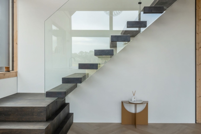 Minimalistisch interieur met moderne, zwevende trap en glazen leuningen in gezinshuis. Donkere treden en houten tafel met wit decoratief item. Grote ramen zorgen voor veel natuurlijk licht. - Finnhouse