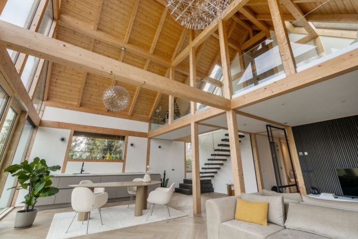 Ruime moderne woonkamer met hoge houten plafonds, grote ramen en open indeling, ideaal voor een gezin. Beige bank, eettafel met stoelen, kamerplanten en trap naar bovenverdieping. Neutrale tinten zorgen voor warme sfeer. - Finnhouse