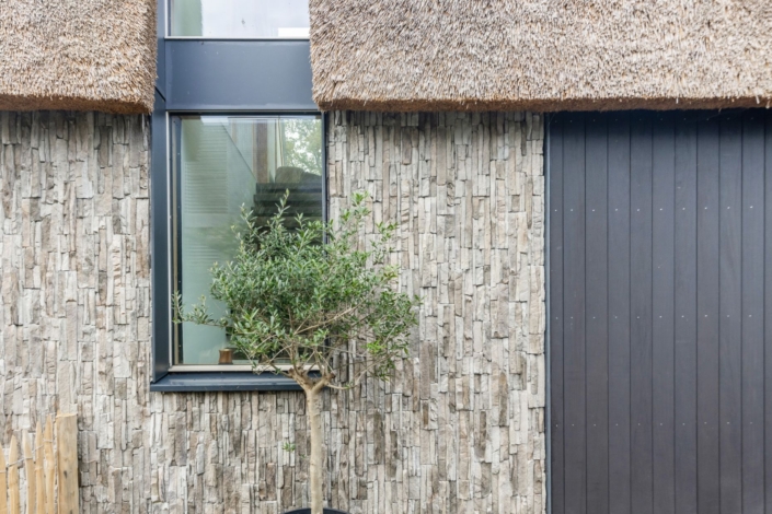 Een moderne voorgevel verticale verticale donkere houten latten met stenen tegels. Een olijfboom in pot voor een groot raam gezelligheid toe, met een charmant rieten dak erboven. - Finnhouse