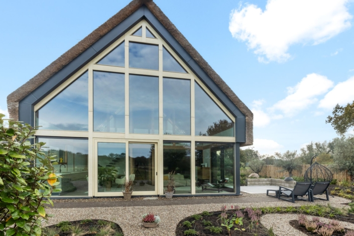 Modern huis met grote glazen ramen en rieten dak, ideaal voor een gezin. De aangelegde tuin heeft grindpaden en planten. Twee ligstoelen op het terras onder een deels bewolkte blauwe lucht. - Finnhouse