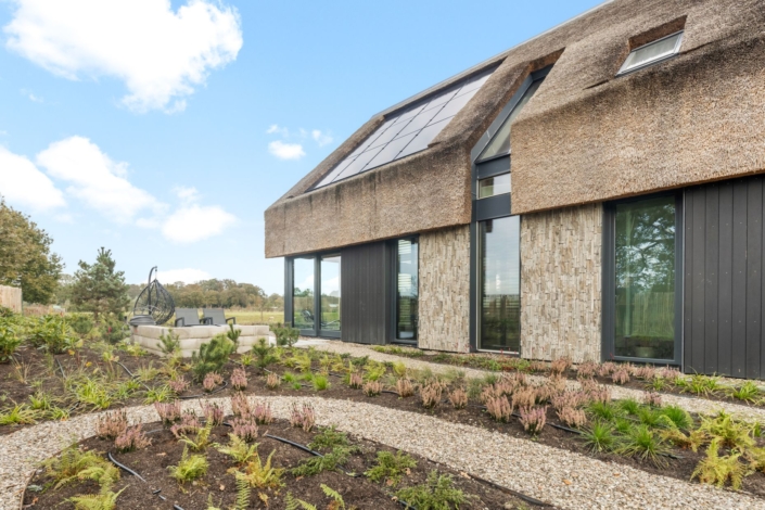 Moderne vakantiewoning met rieten dak en zonnepanelen, omringd door een verzorgde tuin met grindpad naar uitnodigende zitplek. Grote glazen deuren en stenen accenten stralen luxe uit onder een blauwe lucht. - Finnhouse