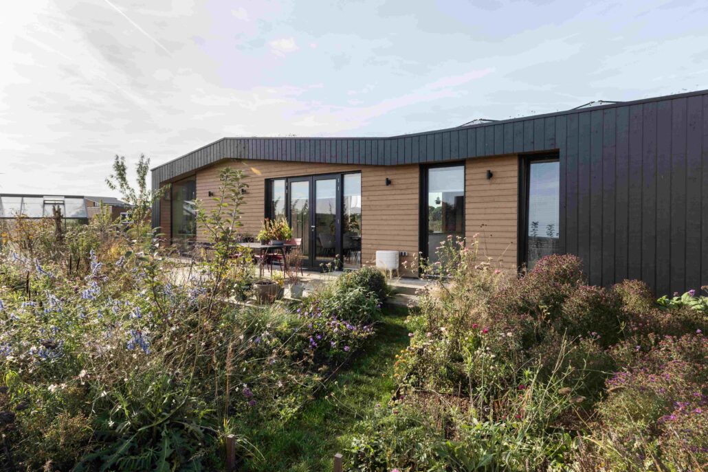 Een modern huis met plat dak en houten bekleding, grote glazen deuren, toont duurzaam bouwen. Voorgrond heeft een vloeibare tuin met diverse planten en bloemen onder een deels bewolkte hemel. - Finnhouse