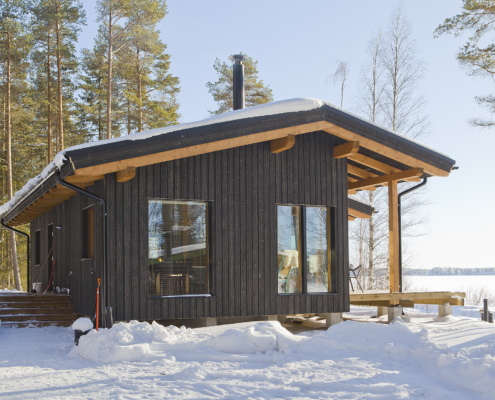 Sneeuw bedekt een zwarte houten Finnhouse-blokhut tussen hoge dennenbomen, grote ramen weerspiegelen de blauwe lucht. Op de achtergrond een bevroren meer en besneeuwde bomen in het winterlandschap. - Finnhouse
