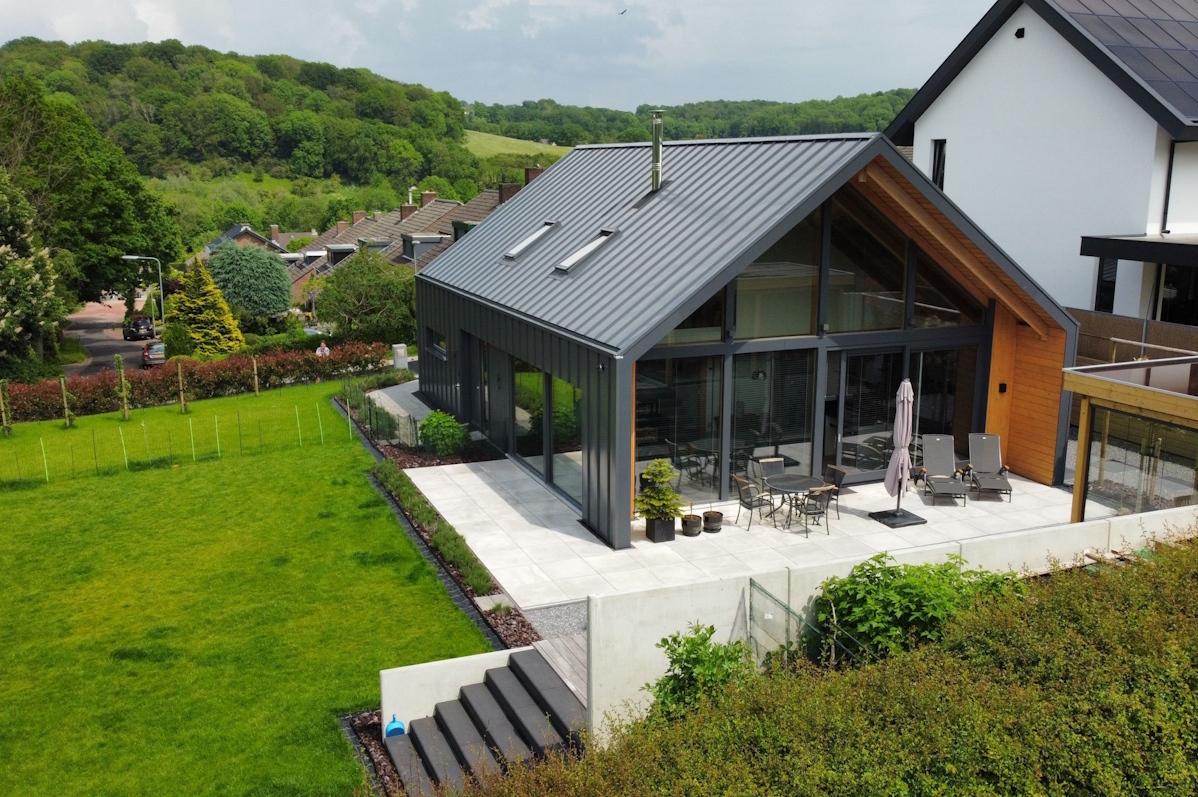 Moderne schuurwoning met schuine metalen dak en grote glazen ramen, gelegen op een heuvel met ruim terras en tuin, daarnaast door groen en uitzicht op andere huizen en beboste heuvels. - Finnhouse