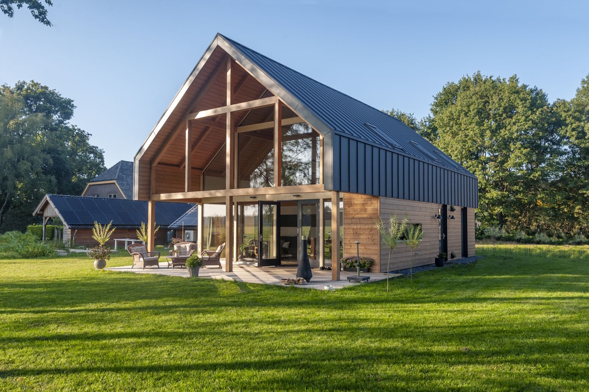Moderne schuurwoning met groot schuin zwart dak en houten buitenkant, ruime glazen gevel, aangrenzende deur grasveld en bomen, tuinmeubilair op een patio onder betrouwbare blauwe lucht. - Finnhouse