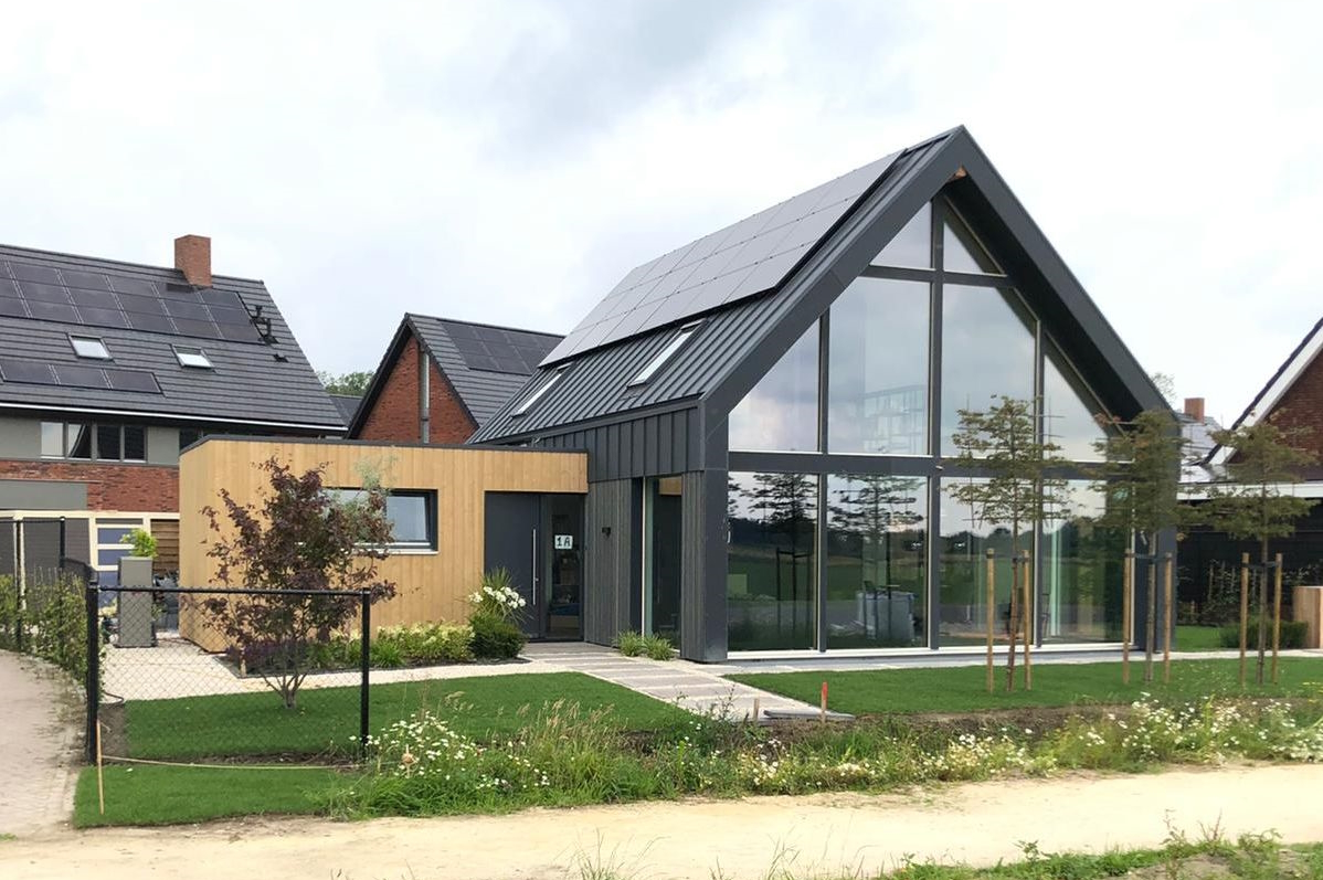 Moderne schuurwoning met zadeldak en grote glazen gevel, houten en donkere metalen bekleding, voorzien van zonnepanelen. Voortuin bevat kleine bomen en pad naar de uitnodigende ingang. - Finnhouse