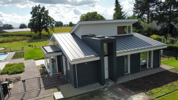 Moderne bungalow met strak, donkergrijs metalen dak en witte muren, daarnaast door goed onderhouden tuin en ruim grasveld. Zonovergoten dag met blauwe lucht en verspreide wolken zichtbaar de serene sfeer. - Finnhouse