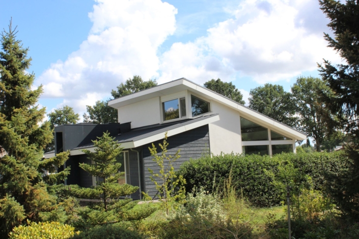 Moderne Finnhouse bungalow met schuin dak, grote ramen en witte en donkergrijze gevelbekleding. Omgeven door dubbele bomen en struiken onder een blauwe hemel met wolken. - Finnhouse