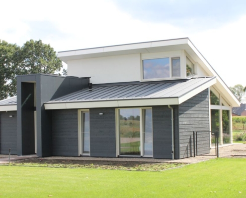 Moderne Finnhouse bungalow met donkergrijze en witte buitenkant, grote ramen en schuin dak. Voortuin met strak gemaaid gras, omlijst door bomen en een veld op de achtergrond. - Finnhouse