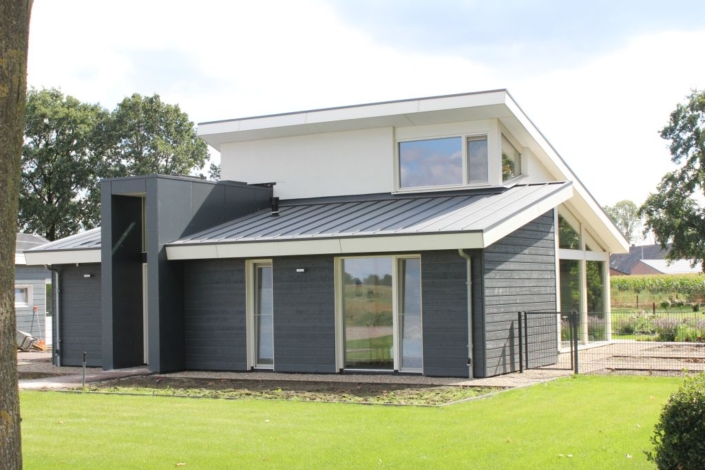 Moderne Finnhouse bungalow met donkergrijze en witte buitenkant, grote ramen en schuin dak. Voortuin met strak gemaaid gras, omlijst door bomen en een veld op de achtergrond. - Finnhouse
