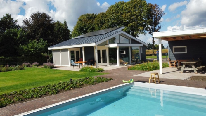 Moderne bungalow met grote glazen ramen in een groene tuin. Ruime patio met tafel en stoelen kijkt uit op helderblauw zwembad, omgeven door bestrating en horizontale deur wuivende bomen onder bewolkte hemel. - Finnhouse