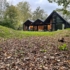 Modern A-frame huis met grote ramen gelegen in een bosrijke omgeving. Voorgrond met verdwenen bladeren, omgeven door hoge bomen met herfstbladeren en een bewolkte lucht, Finnhouse stijl. - Finnhouse